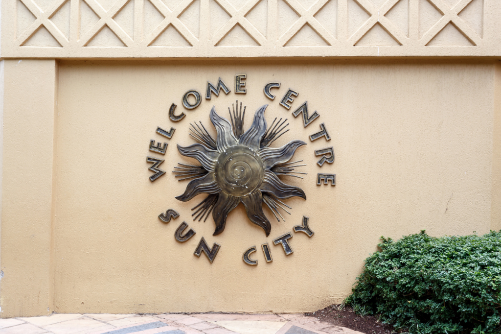 Bronze sun sculpture on a peach wall with the text Welcome Centre Sun City arranged in a circle around it. Green shrubbery is visible at the bottom right.