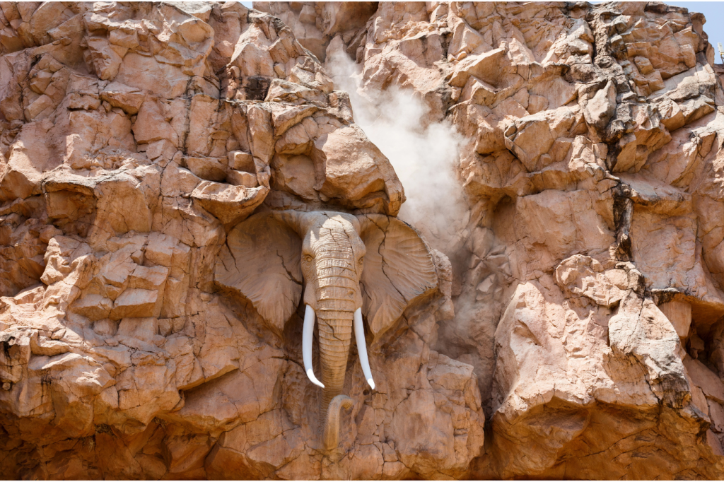 A large stone sculpture of an elephant head with tusks is carved into a rocky cliff face. Dust surrounds the sculpture, suggesting recent carving or erosion, with the sun illuminating the textured rocks.