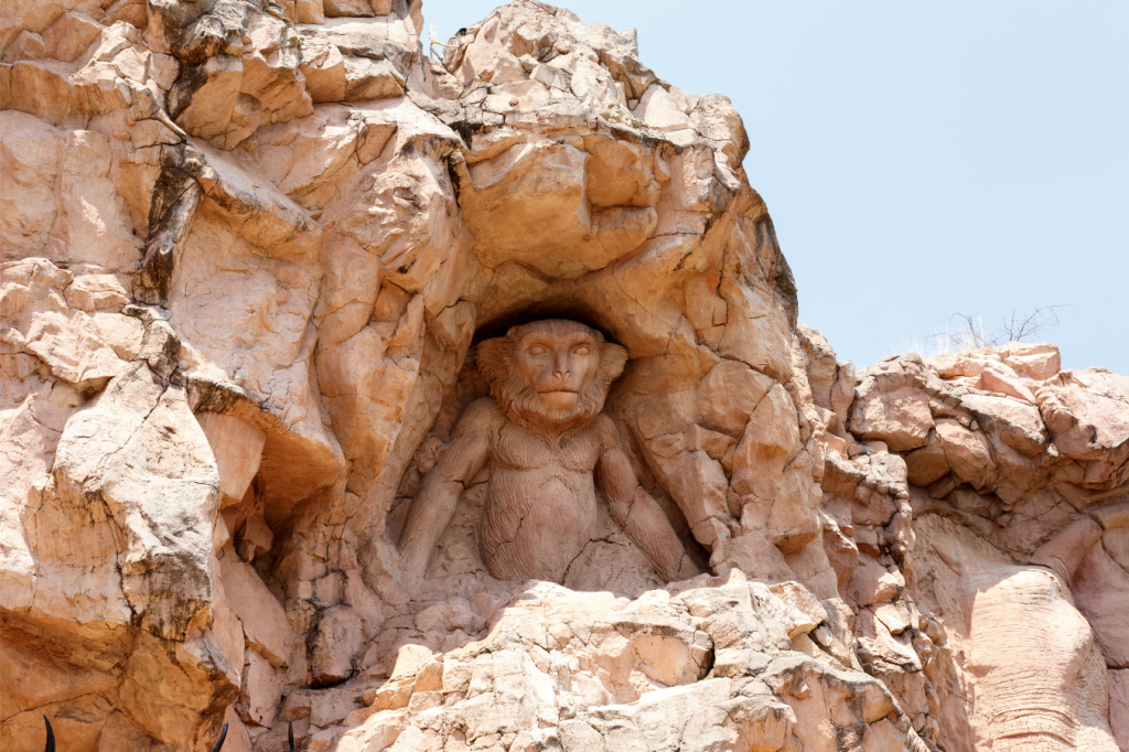 A large stone sculpture of a seated monkey is carved into a rocky cliff under a clear sky. The intricate details showcase the monkeys facial features and limbs, blending naturally with the surrounding rugged rock formations.