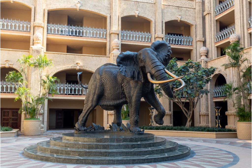 A large elephant statue stands on a circular stone pedestal in an open courtyard. The courtyard is surrounded by a beige multi-story building with ornate railings and arches. Lush potted plants are placed around the base of the statue.