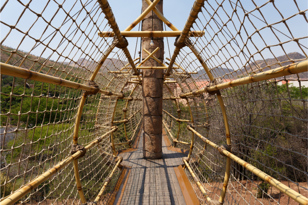 A narrow bridge made of wooden planks and ropes stretches across a canopy of green trees. The ropes form a protective netting on both sides. Distant mountains are visible under a clear blue sky.