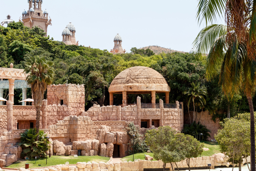 A large, domed stone structure surrounded by lush greenery and palm trees under a clear sky. The scene resembles a historical or ancient architectural site with towers visible in the background.