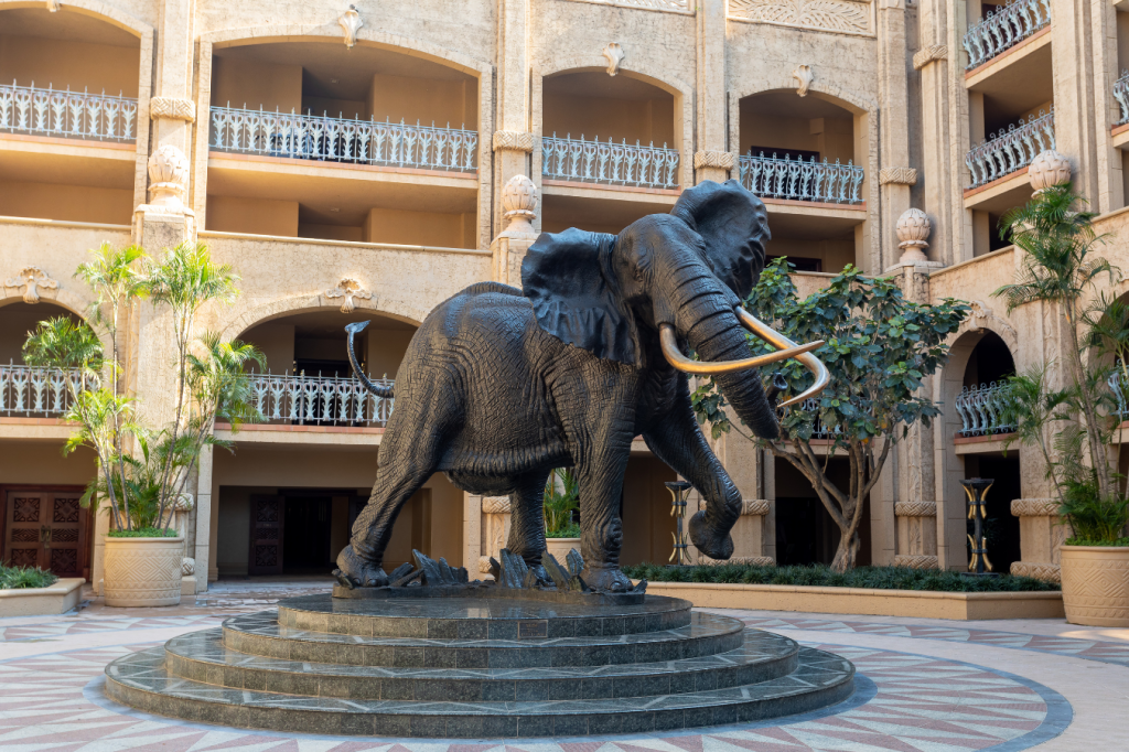 A large elephant statue stands on a circular platform in a grand courtyard. The ornate building with decorative balconies and lush green plants surrounds the statue, creating an opulent and serene atmosphere.