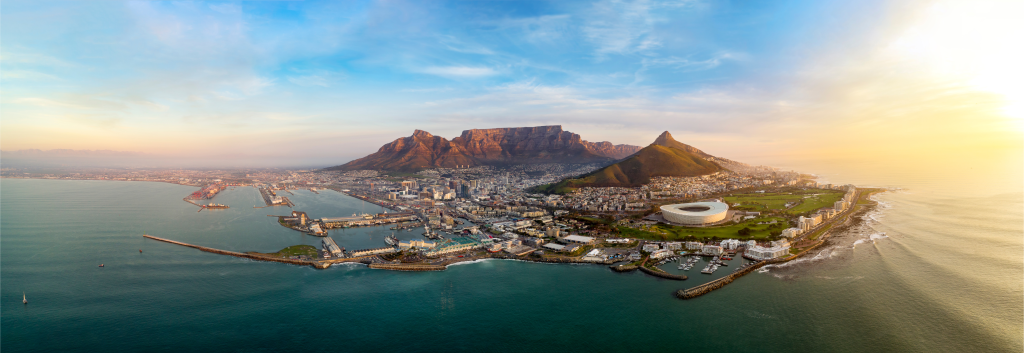 Aerial view of a coastal city with a prominent stadium, surrounded by water. A mountain range forms a dramatic backdrop under a clear blue sky, transitioning to a warm sunset on the horizon.