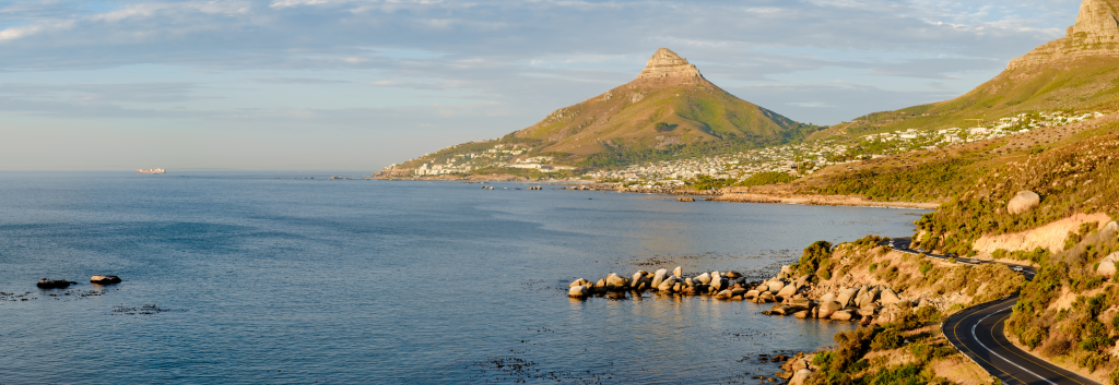 A scenic coastal view shows a curving road alongside a calm ocean. Rugged mountains rise in the background, covered with green vegetation, while a cluster of white buildings nestles at the mountains base near the shoreline.