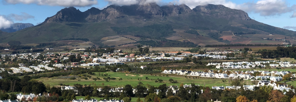 A scenic view of a town with clusters of white houses, surrounded by lush greenery and open fields. In the background, a range of majestic mountains is partially covered with clouds under a blue sky.