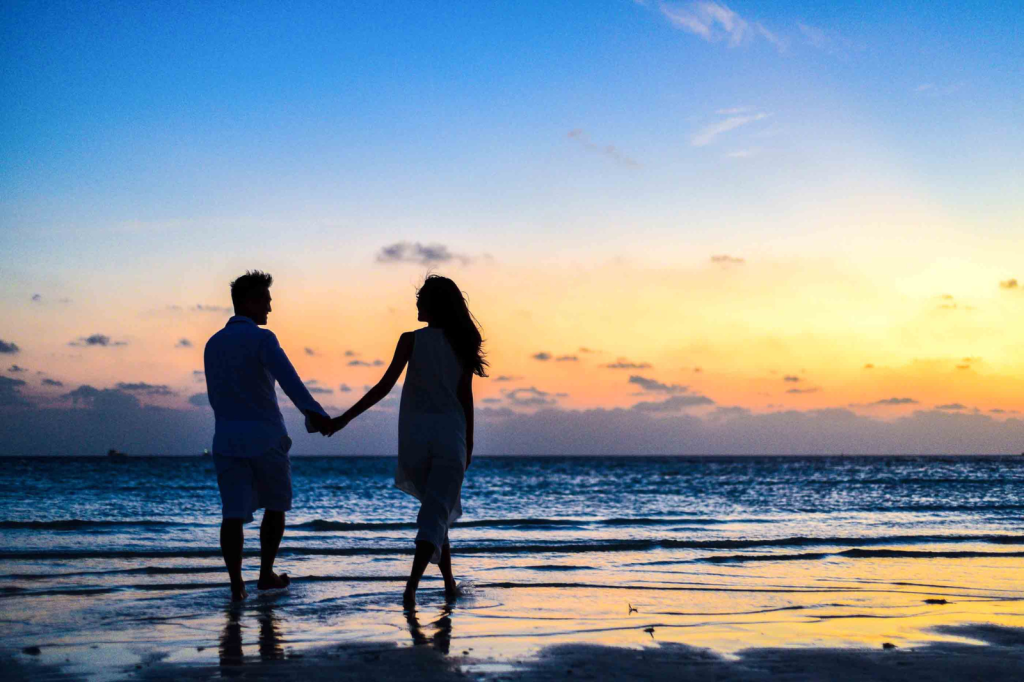 A couple walks hand in hand along the beach at sunset. The sky is a blend of blue, orange, and yellow hues, reflecting on the calm water. Their silhouettes are visible against the vibrant sky.