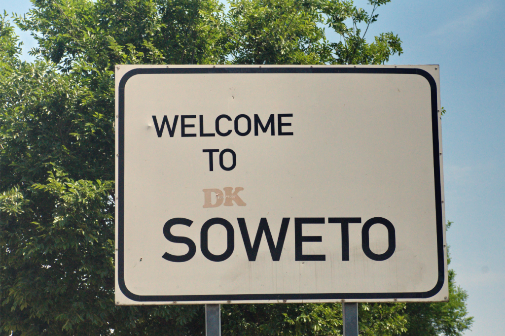 A white sign with black text reads Welcome to Soweto against a backdrop of green trees and a blue sky.