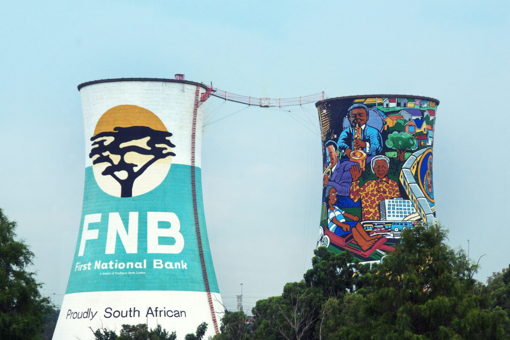 Two colorful cooling towers, one with the FNB logo and Proudly South African text, the other featuring a vibrant mural of South African cultural scenes. Greenery is visible in the foreground.