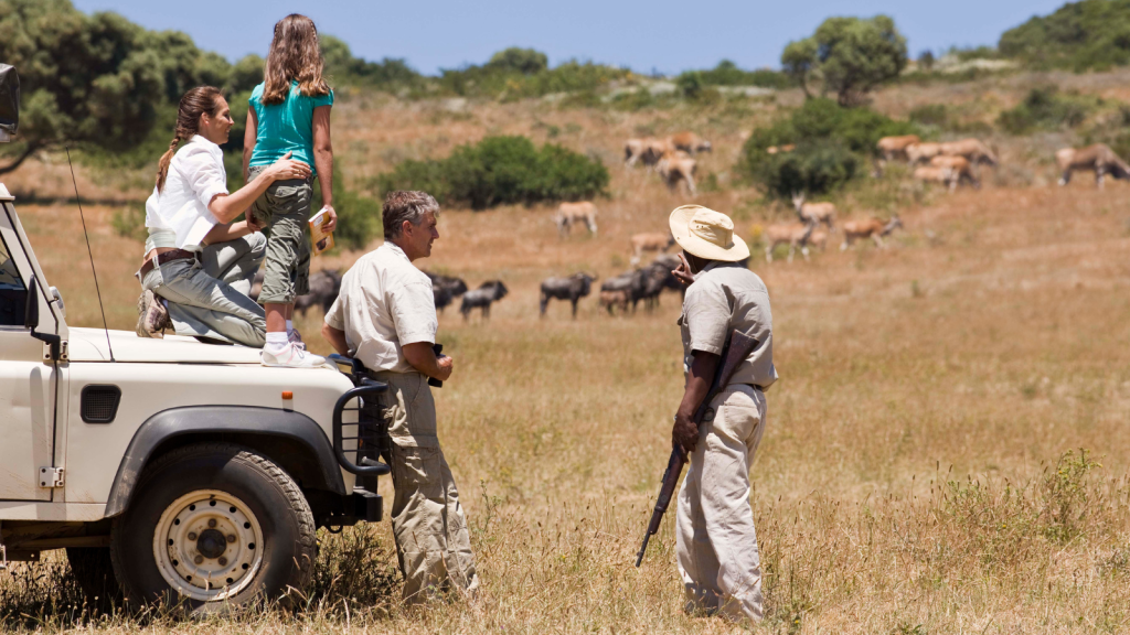 A woman, a girl, and two men stand near a safari vehicle in a grassy landscape. The woman and girl observe wildlife in the distance, while one man holds a camera and the other, wearing a hat, stands near the vehicle. Animals are grazing in the background.