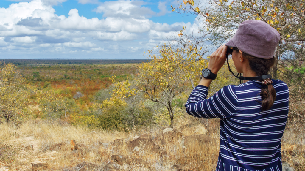 A person wearing a striped shirt and a hat looks through binoculars at a vast landscape. The scene features dry grass, scattered trees, and a wide horizon under a partly cloudy sky.