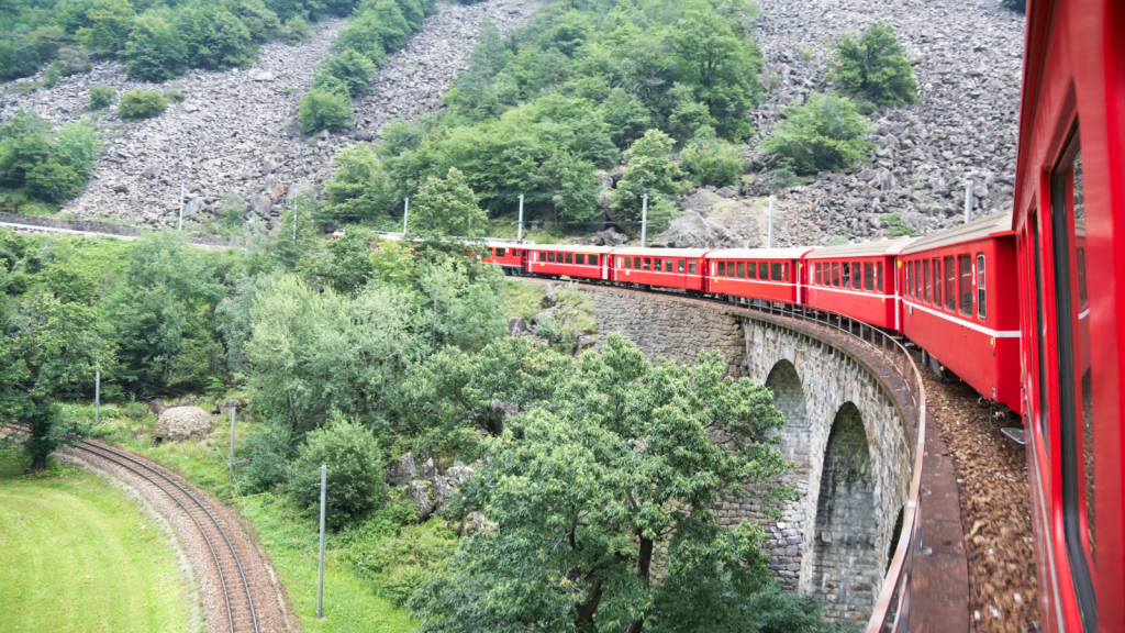 A bright red train crosses a stone bridge amidst lush green scenery. The train curves around the hillside, with trees and rocky terrain visible. The view captures the dynamic movement of the train through the picturesque landscape.