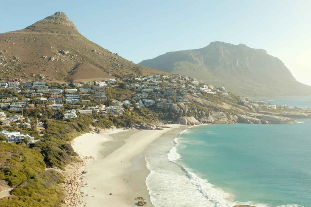 A coastal landscape featuring a sandy beach with gentle waves, nestled alongside a rocky shoreline. Houses are scattered across the hillside, with two prominent mountains in the background under a clear blue sky.