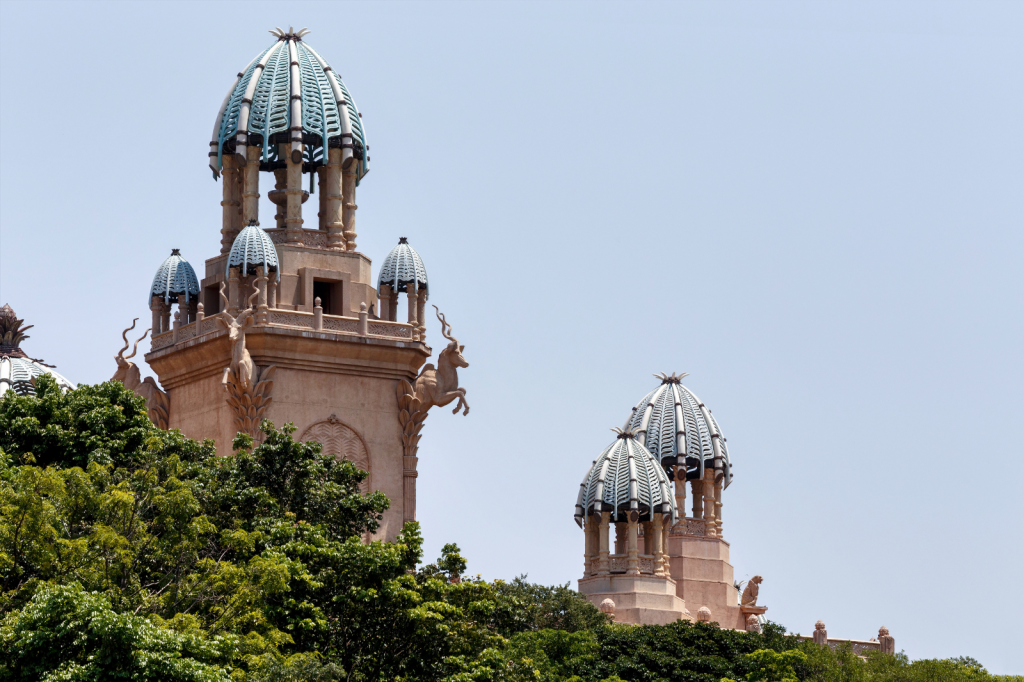 The image shows a large architectural structure with ornate, dome-shaped spires, resembling an exotic palace. The building is surrounded by lush green trees under a clear blue sky.