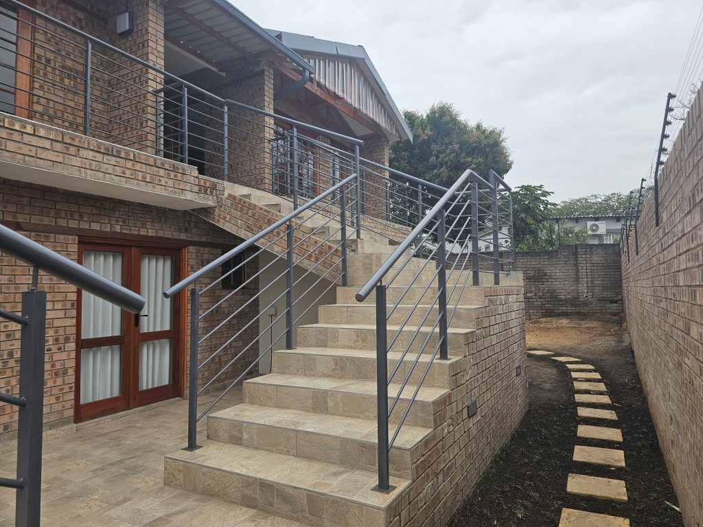 A modern brick building with an exterior staircase leading to the second floor. The staircase has metal railings and beige steps. A paved path runs alongside a brick wall and leads to a garden area with trees. The sky is cloudy.