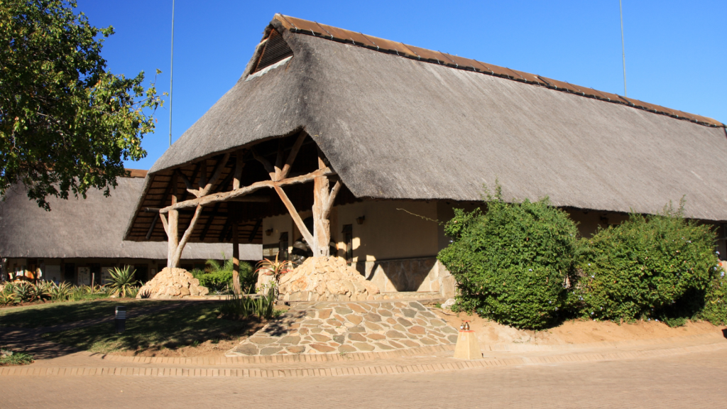 A rustic building with a thatched roof and wooden beams, partially surrounded by lush greenery, under a clear blue sky. The structure appears to be situated in a warm, sunny location, with a stone path leading to the entryway.