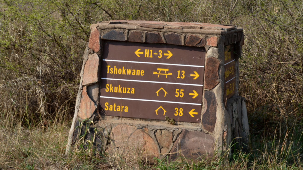 A stone signpost in a grassy area displays directions and distances: Tshokwane (13 km), Skukuza (55 km), and Satara (38 km) with arrows pointing right or straight on a brown background.