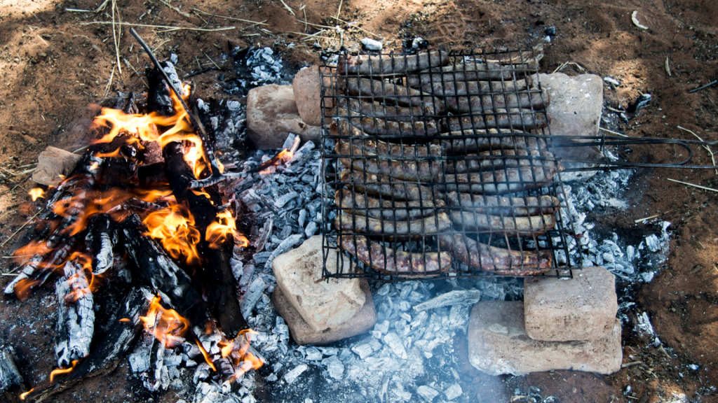 Sausages are being grilled on a metal rack over an open fire, surrounded by bricks. The flames and coals are visible on the left, and smoke is rising from the sausages as they cook. The ground is earthy, with some dry grass.