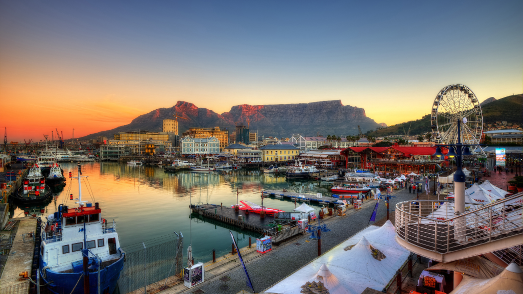 Scenic view of a waterfront harbor at sunset, featuring boats docked near the vibrant waterfront buildings. A large ferris wheel stands on the right, with a mountainous backdrop under a colorful sky.