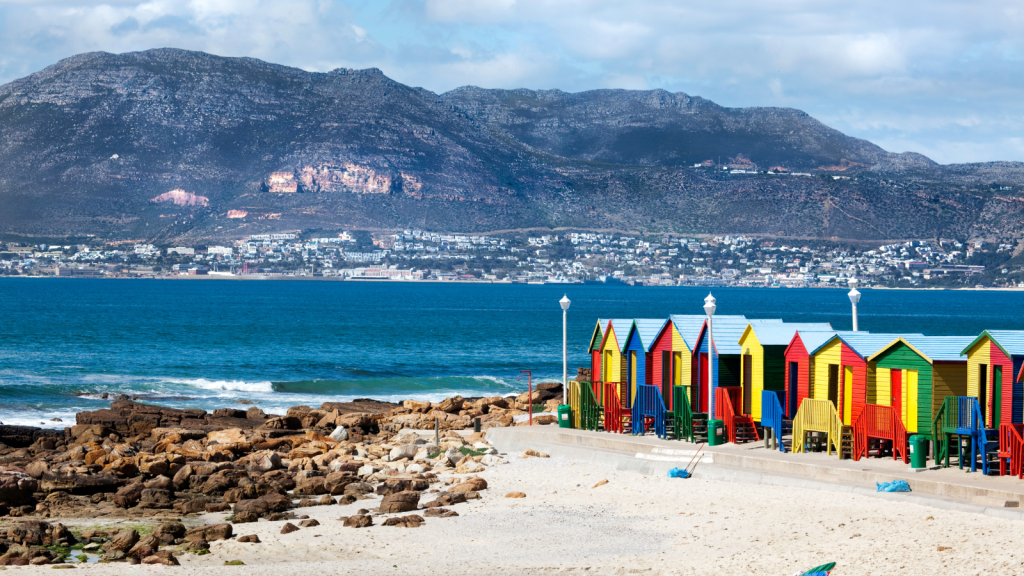 Colorful beach huts line a sandy shore with rocky outcrops, set against a backdrop of blue ocean and distant hills under a partly cloudy sky.