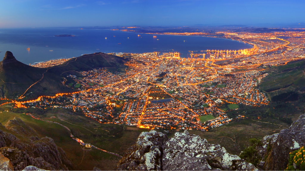 Panoramic view of a city illuminated at night, bordered by a coastline. The city lights form a network of bright patterns against a dark landscape, with a large mountain and ocean creating a picturesque backdrop.