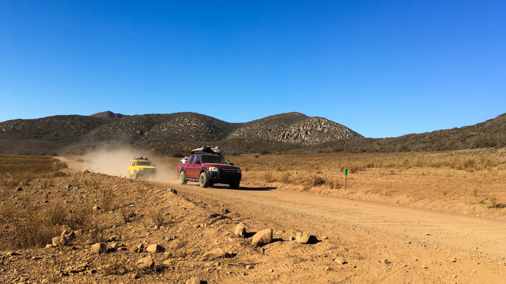 Two off-road vehicles drive on a dusty dirt road in a dry, rocky landscape. The lead vehicle is red, followed by a yellow one, with hills and clear blue skies in the background. Dust trails behind the vehicles.