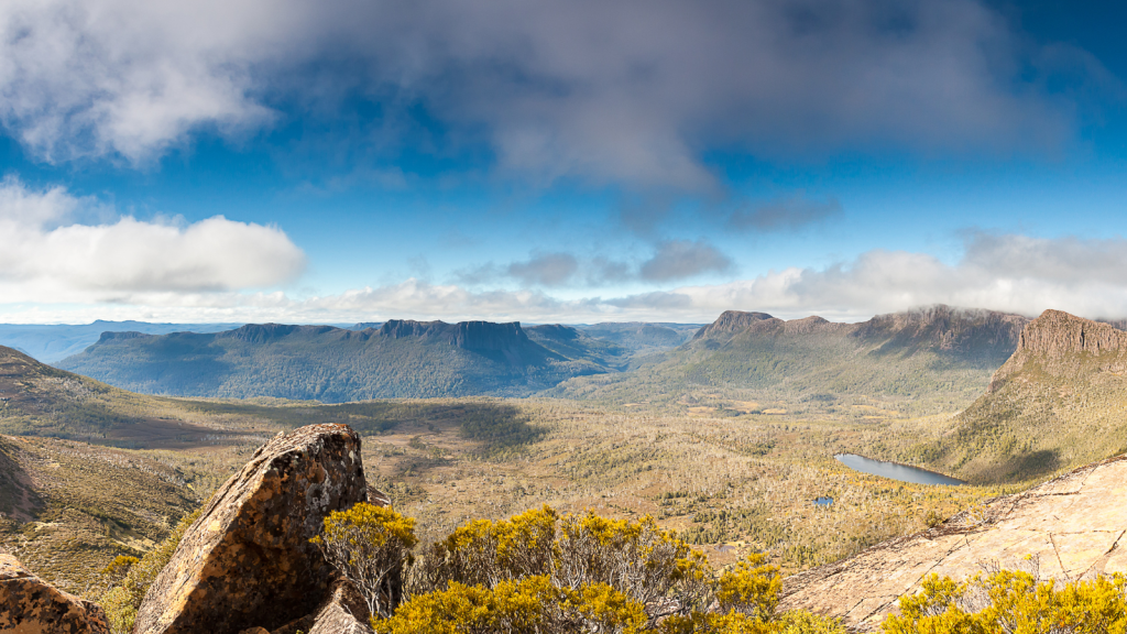 Panoramic view of a vast, rugged landscape featuring rocky formations, a small lake, and sprawling greenery under a partly cloudy blue sky. Distant mountain ranges form the backdrop of the scenic terrain.