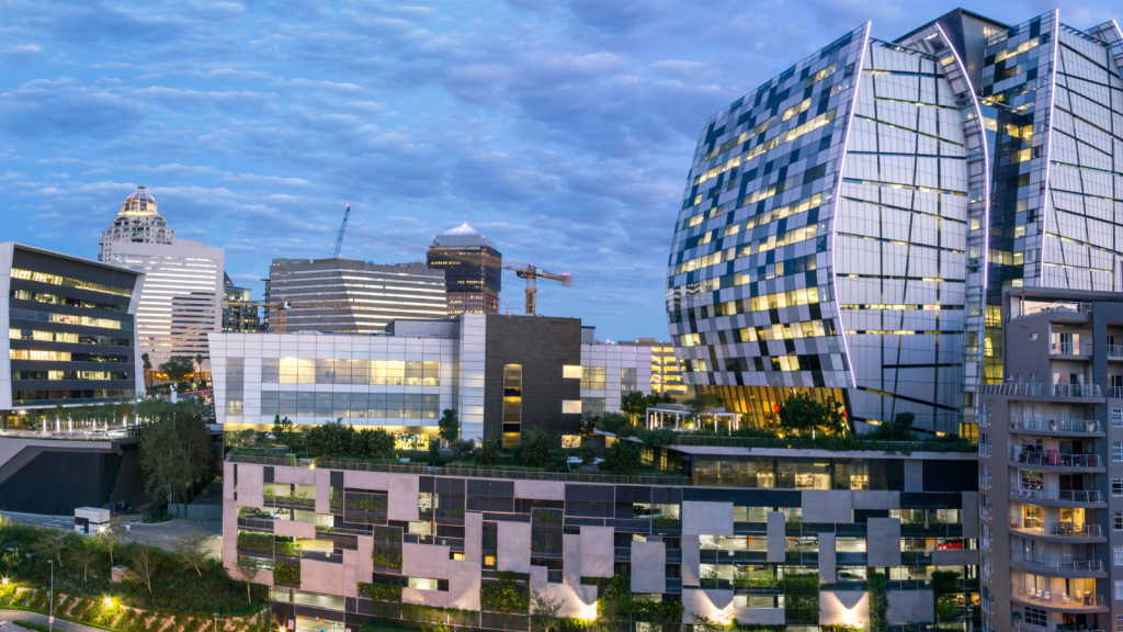 A contemporary cityscape at dusk featuring modern office buildings with unique geometric designs and glass facades. A construction crane is visible amidst other skyscrapers under a partly cloudy sky.