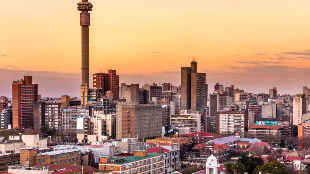 A panoramic view of Johannesburgs cityscape at sunset, featuring a tall telecommunications tower and a variety of modern and older buildings under a colorful sky.