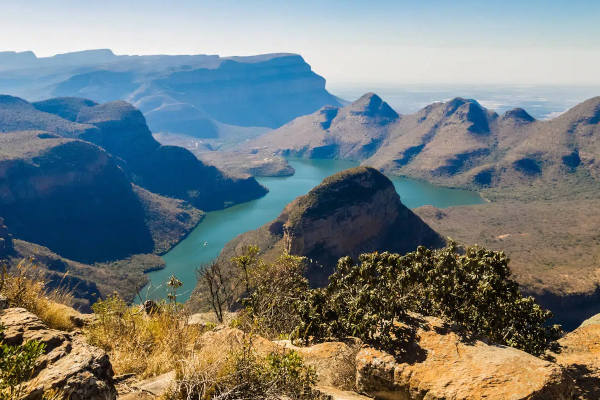 A scenic view of Blyde River Canyon in South Africa, featuring lush green vegetation, rocky cliffs, and a winding river. The canyon stretches into the distance with a clear blue sky above.