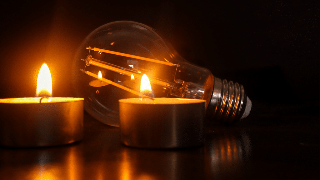 Two lit candles placed in front of an unlit light bulb, set against a dark background. The warm glow of the candles contrasts with the cool metallic surface of the bulb, creating a serene and contemplative atmosphere.