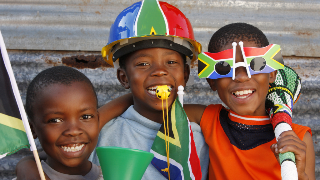 Three children smile while celebrating, wearing colorful hats and glasses with South African flags, holding matching flags and a vuvuzela. They stand close together against a metallic background, showing joy and excitement.