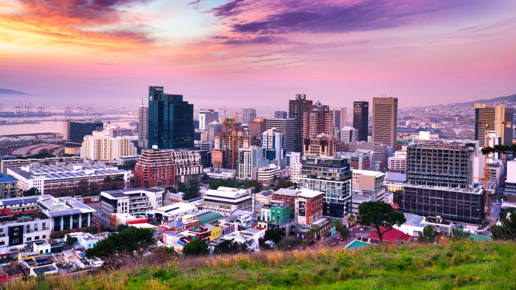 A vibrant cityscape at sunset with a mix of modern skyscrapers and colorful buildings. The sky is painted in shades of purple, pink, and orange, while greenery in the foreground adds contrast to the urban landscape.