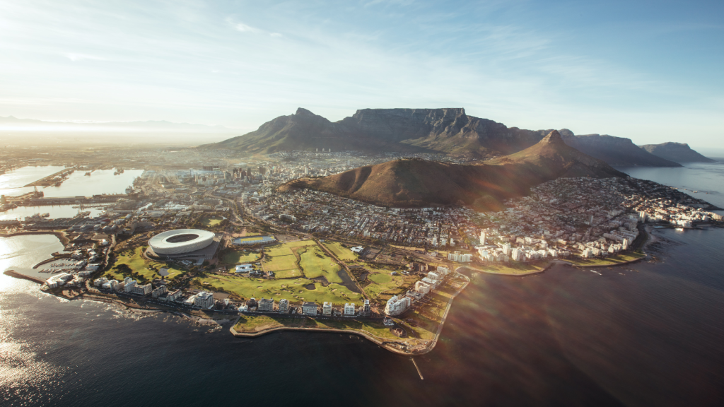 Aerial view of Cape Town, South Africa, featuring the coastline, green fields, and the prominent Cape Town Stadium. Table Mountain looms in the background under a clear sky, with the city extending along the shoreline.