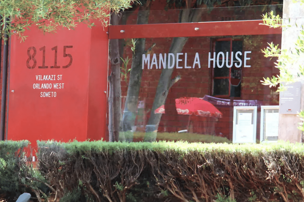 Red building exterior with 8115 Vilakazi St, Orlando West, Soweto and Mandela House written on it. The entrance is surrounded by bushes, with a glass window reflecting trees and red umbrellas.