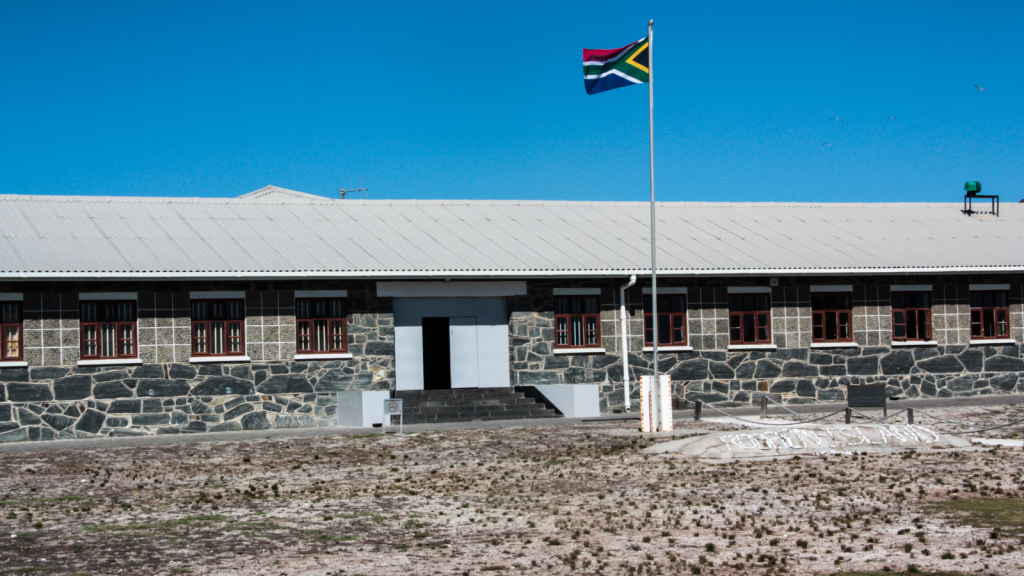 A stone building with a corrugated metal roof sits under a clear blue sky. A South African flag flies on a pole in front of the building. The ground is dry and barren, with sparse vegetation.