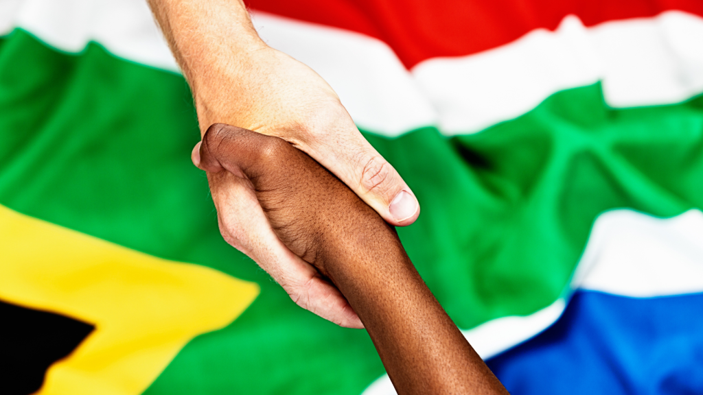 Close-up of two hands shaking in front of a blurred South African flag. One hand is light-skinned and the other is dark-skinned, symbolizing unity and diversity.