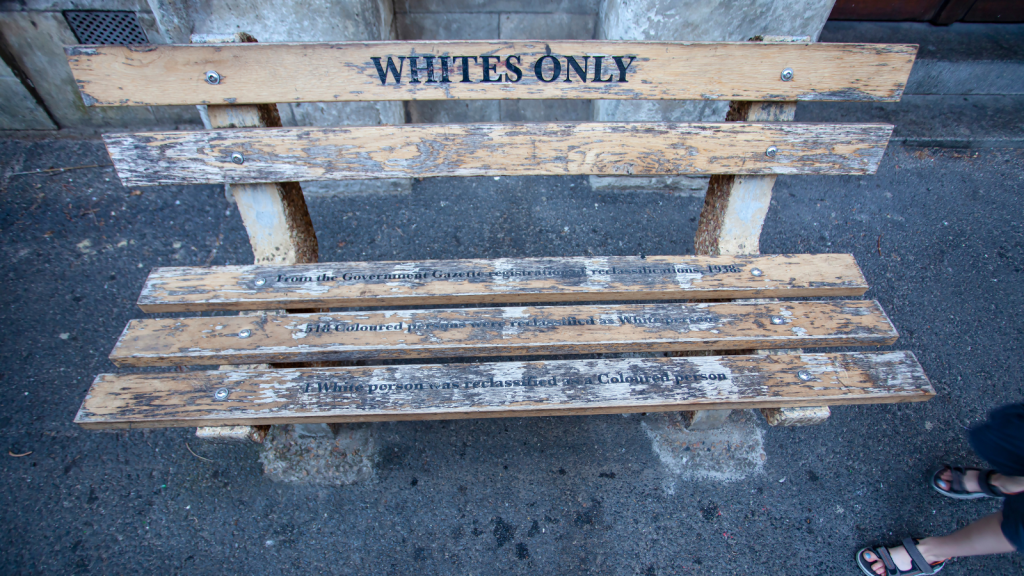 A weathered wooden bench with WHITES ONLY painted on the backrest. Additional faded text regarding segregation policies is visible on the seat slats. The scene includes part of a persons leg near the bench.