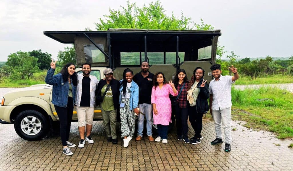 A group of people smiling and posing in front of a safari vehicle on a paved area. The background features green trees and a cloudy sky. Some individuals are making peace signs with their hands.