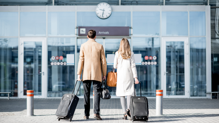 A man and a woman walk towards an airport arrivals entrance, each pulling a suitcase. They are wearing long coats. A large clock is visible above the entrance doors.