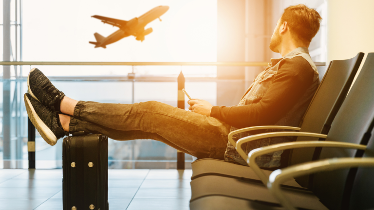 A man sits in an airport terminal with his legs stretched out on a suitcase, holding a smartphone. He gazes out the window at an airplane taking off against a bright sky.