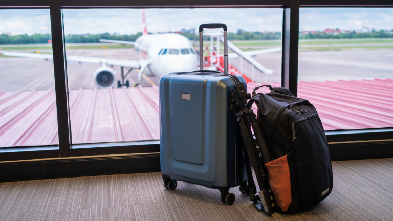 A blue suitcase and a black backpack are placed on the floor in an airport terminal, with a large window showing an airplane parked on the runway outside. The sky is partly cloudy, and the scene conveys a travel atmosphere.