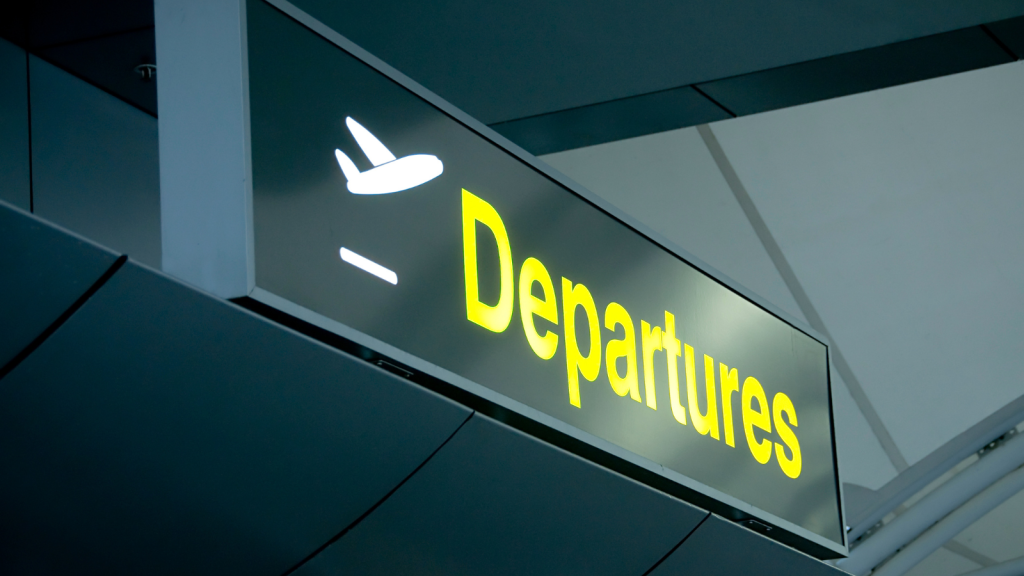 A close-up of an airport sign displaying the word Departures in large yellow letters and an airplane icon, indicating the area for outgoing flights. The sign is mounted on a modern architectural structure.