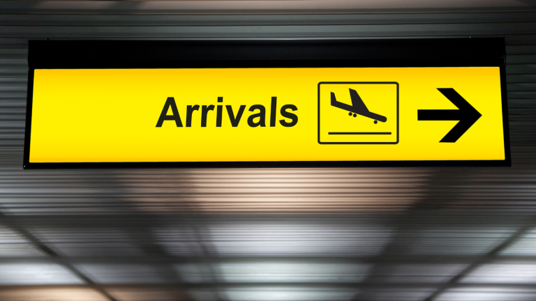 A bright yellow airport sign with Arrivals in black text and an airplane icon, directing travelers with a black arrow to the right. The background shows a blurred airport ceiling.