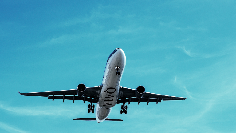 A large airplane with Qatar on the side is flying through a clear blue sky. The plane is captured from below, showing its wings extended and landing gear visible.