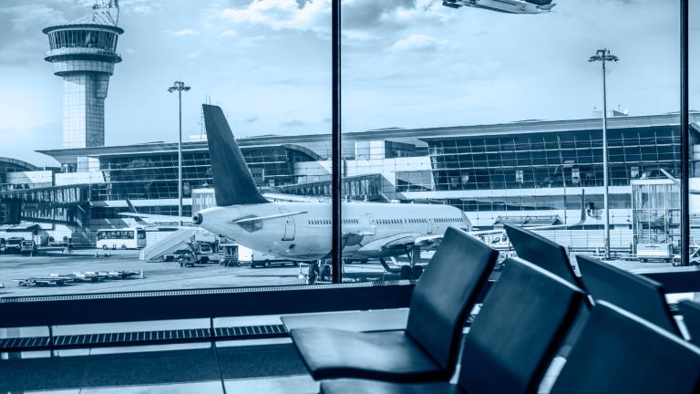 An airport scene with a passenger airplane parked at the gate. The terminal building and control tower are visible in the background, and empty seats are in the foreground, viewed from inside the terminal through large windows.