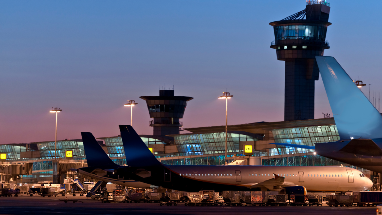 A commercial airplane is parked at an airport terminal during twilight. Several control towers are visible in the background, and the sky is a blend of deep blue and warm hues. The scene is illuminated by ambient and artificial lighting.