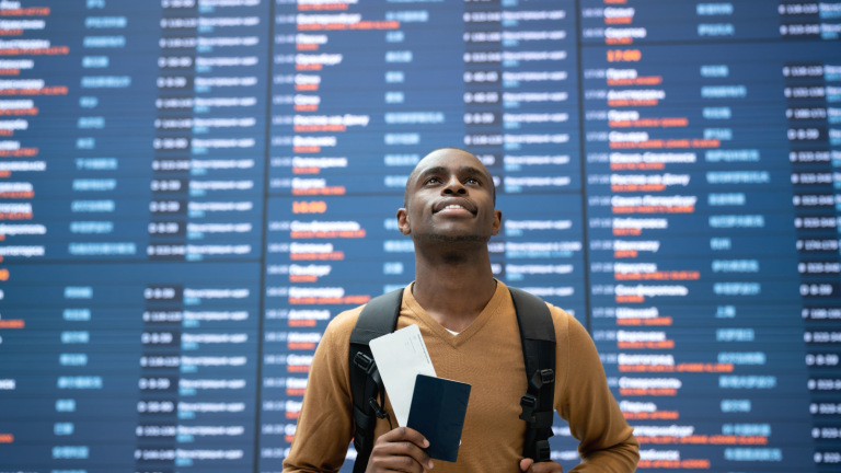 A man stands in front of a large airport departure board, looking up. Hes holding a passport and boarding pass and has a backpack on. The board displays various flight times and destinations.