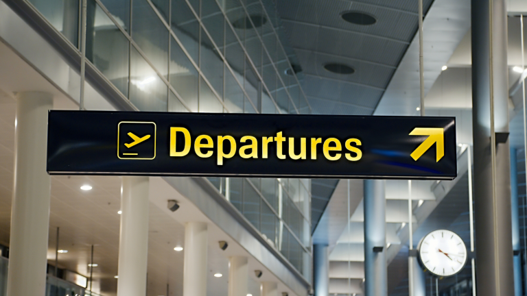 Sign with Departures and an arrow pointing to the right hangs in a modern airport terminal. The background features glass, steel elements, and a visible clock showing 10:08.