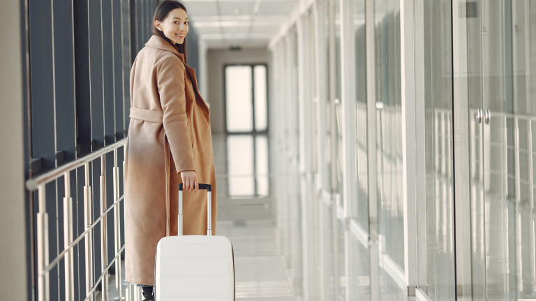 A person in a long coat is pulling a white suitcase through a bright, glass-walled corridor. They are looking back towards the camera with a slight smile.